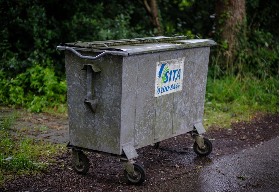 A weathered grey metal waste bin with a partially open lid, situated on a damp, muddy pathway bordered by lush green grass and hedge plants. The bin has four small wheels, with one slightly tilted, an