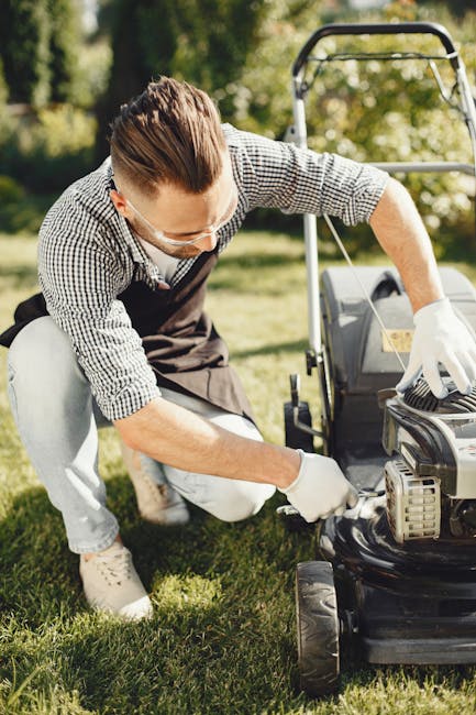 A man wearing safety glasses, gloves, a checkered shirt, and an apron is kneeling on a well-maintained green lawn, inspecting or repairing a lawnmower. The garden features a neatly trimmed hedge and a